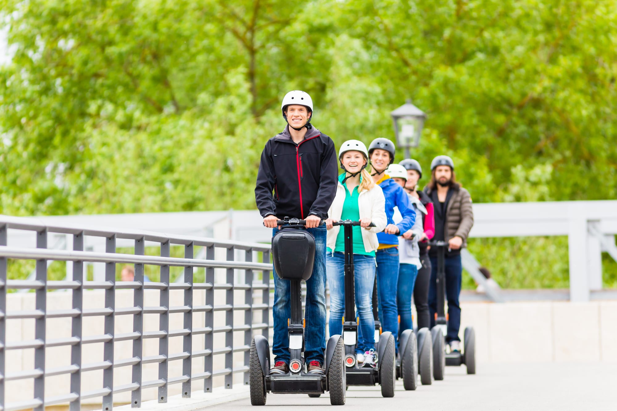 A group of six people wearing helmets and riding segways in a single-file line with green trees in the background.
